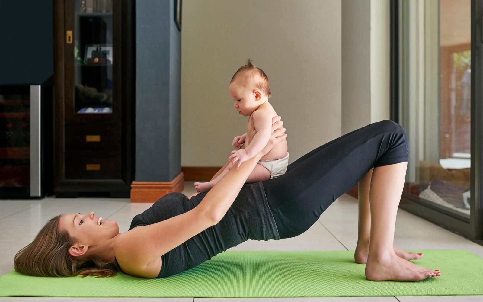 Madre practicando yoga posparto en casa mientras sostiene a su bebé
