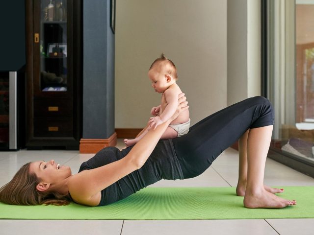 Madre practicando yoga posparto en casa mientras sostiene a su bebé